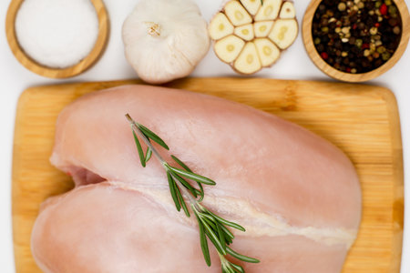 A close-up view from above of chicken breast fillet with a sprig of rosemary lying on a wooden board, nearby are wooden bowls with pepper, seasoning and salt and garlic on a white background. The concept of dietary dishes, a recipe for healthy and healthy food. High quality photoの写真素材