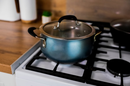 On a white gas stove with a black cast iron grate there is a blue pot with a glass lid and rubber pads on the handles against the background of the kitchen in out-of-focus, Concept of cooking, cooking, cooking. High quality photoの写真素材