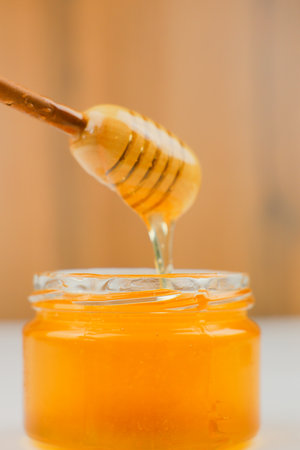 Close-up of a glass jar of liquid yellow honey standing on a white table and into which a wooden spoon is dipped, from which honey drips. The concept of healthy eating, proper and healthy food, natural desserts. High quality photoの写真素材