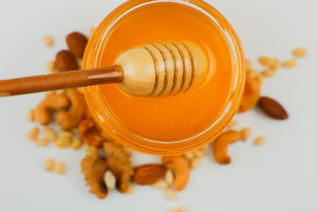 Close-up view from above: a glass jar with liquid yellow honey stands on a white background surrounded by a mix of nuts, a wooden spoon lies on top of it, honey drips from it. The concept of healthy eating, organic and healthy desserts. High quality photoの写真素材