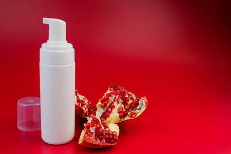 Close-up white foam cleanser with dispenser and open lid to nourish and moisturize the skin stands next to an open pomegranate and pomegranate seeds on a pink background with space for text. The concept of body and face care, skin care, cosmetics with pomegranate extract. High quality photoの写真素材
