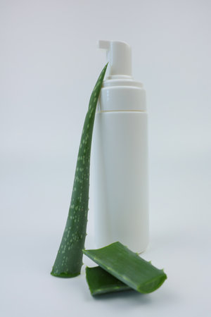 Close-up of a white foam cleanser with a dispenser and an open lid to nourish and moisturize the skin stands on a white background, aloe leaves lie nearby. Body and face care concept, skin care, cosmetics with aloe extract. High quality photoの写真素材