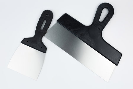 Close-up of two metal spatulas with black handles of different sizes lying on the side on a white...