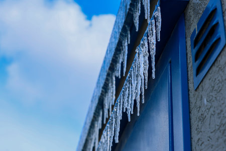 Side view of the blue roof, from which icicles hang in two rows against the background of a blue sky with clouds. The concept of danger, frozen water, winter. High quality photoの写真素材