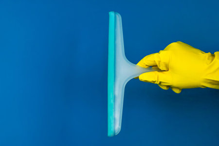 Close-up on the right, a hand wearing a yellow rubber cleaning glove holds a blue cleaning tool on a...
