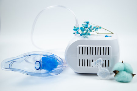 Close-up of the breathing blue mask of the inhaler lying against the background of the machine, flowers and ampoule with medicine out of focus on a white background. The concept of treating respiratory diseases, lung diseases, prevention and self-care. High quality photoの写真素材