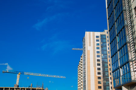 Modern residential buildings are being built with cranes in the foreground against a bright blue sky, highlighting the citys growth and construction activity. High quality photoの写真素材