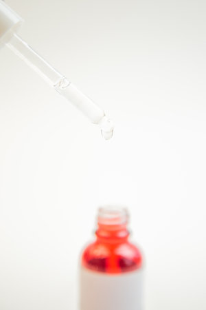 Close-up view of a dropper dispensing liquid above a vibrant red bottle, highlighting the skincare products texture and application in a clean, minimalist environmentの写真素材