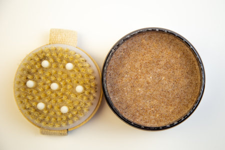 Round container of natural body scrub next to an exfoliating brush, highlighting organic skincare essentials for self-care and pampering experiencesの写真素材