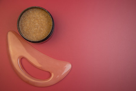 Skincare essentials featuring a jar of exfoliating scrub and a gua sha tool on a bright pink surface, promoting beauty and wellness for self-care practicesの写真素材
