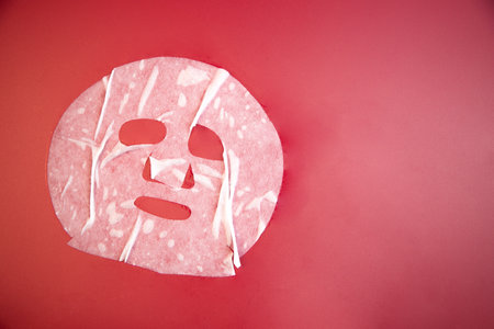 Skincare facial mask sheet with cut-out features is positioned on a bright red background, emphasizing beauty treatment and self-care for relaxation and rejuvenationの写真素材