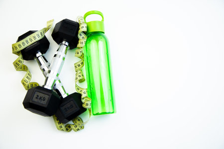 Fitness gear featuring black dumbbells, a vibrant green water bottle, and a measuring tape on a bright white background, representing a healthy lifestyle and exercise motivationの写真素材