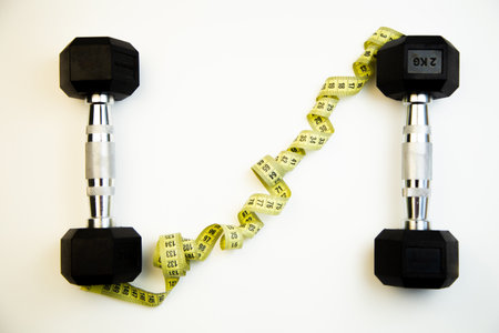 Two black dumbbells are placed on a clean white surface, with a yellow measuring tape artistically intertwined, representing fitness, health, and workout motivationの写真素材