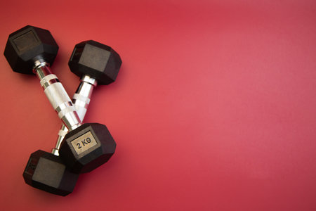 Black dumbbells, each weighing 2 kg, are positioned in a crossed formation on a bright red background, highlighting fitness and strength training essentialsの写真素材
