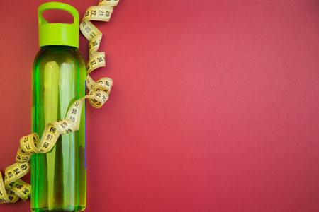 Water bottle in green color, surrounded by yellow measuring tape on a red background, representing hydration and fitness themes for health-conscious individualsの写真素材