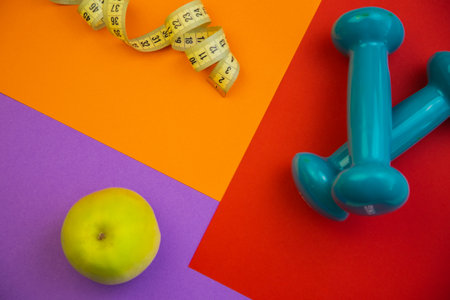 Vibrant flat lay arrangement with green apple, blue dumbbells, and measuring tape on colorful geometric backgrounds, symbolizing health, fitness, and wellness choicesの写真素材