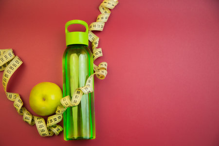 Vibrant green water bottle alongside a yellow apple and measuring tape on a red background, representing health and fitness, ideal for wellness-themed projectsの写真素材