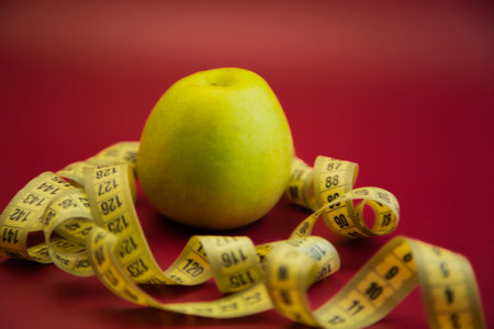 Green apple placed on vibrant red background, surrounded by yellow measuring tape, representing health and nutrition in a creative and colorful arrangementの写真素材