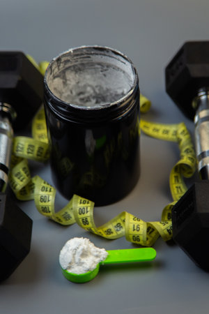 Black protein powder container with green scoop, surrounded by dumbbells and measuring tape,...
