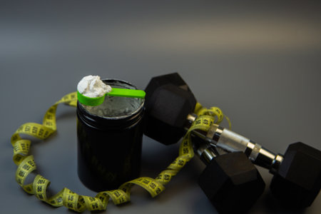 Container of fitness supplement with green scoop, placed beside dumbbells and measuring tape, highlighting the importance of nutrition in strength training and fitness journeyの写真素材
