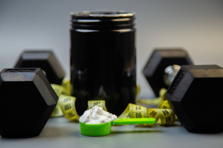 Fitness supplement container with green scoop filled with powder, surrounded by dumbbells, showcasing workout essentials and nutrition for health-conscious individualsの写真素材