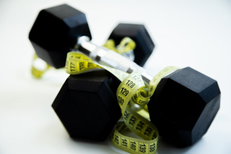 Black dumbbells are intertwined with a yellow measuring tape, representing fitness aspirations and healthy living in a clean, modern workout spaceの写真素材