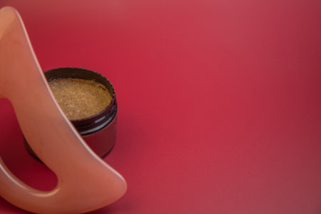 Round jar containing brown sugar scrub is positioned next to a pink applicator on a bright red background, emphasizing beauty and skincare themes with copy spaceの写真素材
