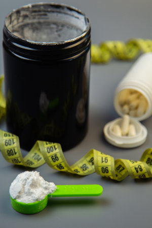 Black supplement container with white powder and pills, green scoop filled with powder, and measuring tape, illustrating health and fitness concepts and choicesの写真素材