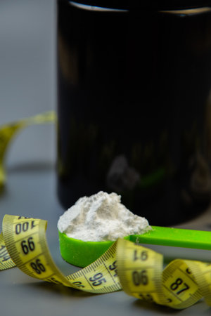Green spoon with white powder is placed in front of a black container, with yellow measuring tape coiled around, emphasizing health, nutrition, and fitness themesの写真素材