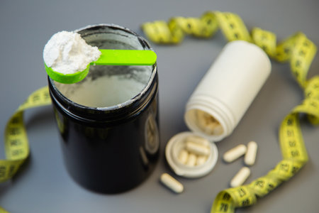 Black jar with white powder and green scoop, next to white pill bottle and scattered capsules, with measuring tape, illustrating health and fitness themesの写真素材