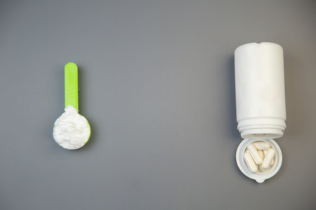 Green measuring spoon with white powder beside a white pill bottle filled with capsules, placed on a smooth gray surface, showcasing health and wellness themesの写真素材