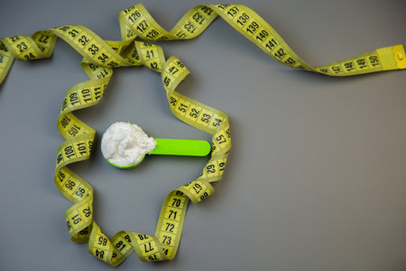 Coiled measuring tape surrounds a green scoop filled with white powder on a gray surface, representing themes of nutrition, fitness, and health awarenessの写真素材