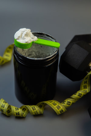 Black protein powder container features green scoop on top, surrounded by yellow measuring tape and...