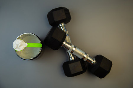Black weights and a protein shake with a green spoon are arranged on a gray background, symbolizing fitness, strength training, and healthy nutrition choicesの写真素材