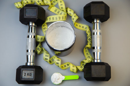 Black dumbbells are positioned alongside a measuring tape and a protein powder container, showcasing fitness essentials for a workout routine and healthy livingの写真素材