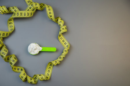 Bright yellow measuring tape forms a circle around a green scoop filled with white powder, representing health and nutrition in a clean, minimalist environment with copy spaceの写真素材