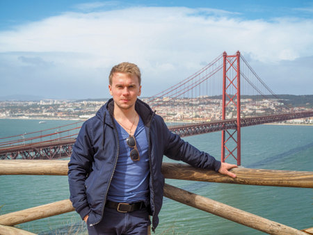 The blonde man stands in the background of the 25th of April bridge in Lisbon. The bridge resembles the Golden bridge in San Franciscoの写真素材