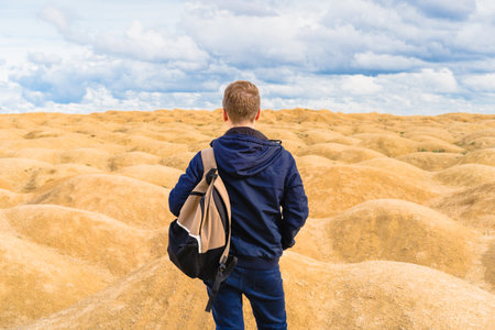 Sporty blond man with a backpack stands with his back to the camera in the desert with sand dunes of...