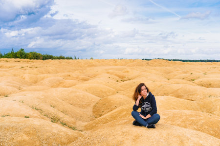 Beautiful young woman in a sweatshirt with an inscription sits on the sand in the desert with sand dunes of bizarre shapeの写真素材