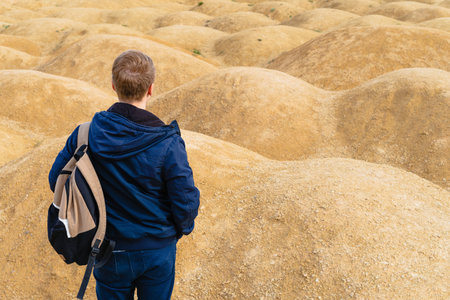 Sporty blond man with a backpack stands with his back to the camera in the desert with sand dunes of bizarre shapeの写真素材