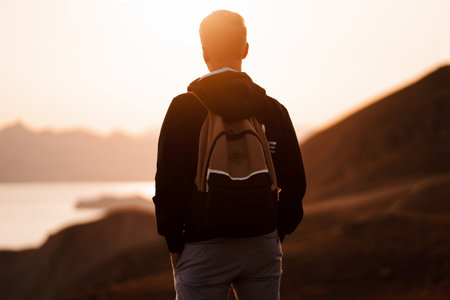 Picturesque mountain landscape at sunset with the sea on the horizon, a young blond man with backpack stands on the slope in the orange rays of the sunsetの写真素材