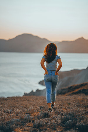 Picturesque mountain landscape at sunset with the sea on the horizon, a young beautiful woman walks down the slope in the orange rays of the sunsetの写真素材