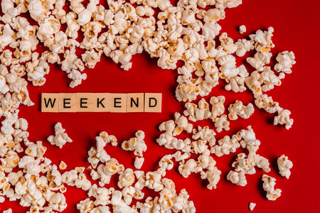 The word "weekend" from wooden blocks lies on a red background surrounded by popcorn. The concept of recreation with watching moviesの写真素材