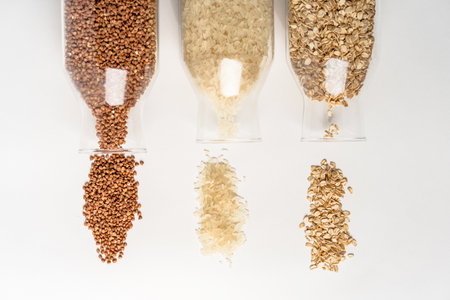 Cereals: buckwheat, rice, oatmeal spilled out of glass containers on a white tableの写真素材