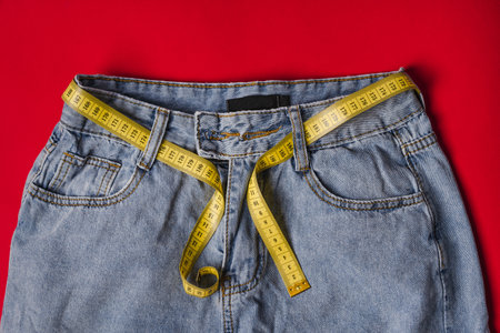 Yellow measuring tape on the waist instead of a belt in jeans on a red background. The concept of...