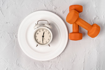 Sports and weight loss concept. The alarm clock is wrapped with measuring tape and dumbbells on a light backgroundの写真素材