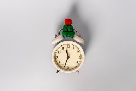 Round alarm clock in a knitted warm green hat on the background. The concept of the winter season....