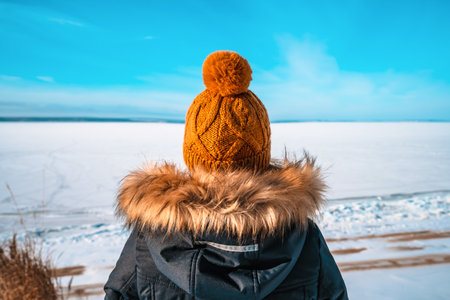 A woman in a winter jacket and a knitted yellow hat stands against the background of a frozen river with a winter landscapeの写真素材