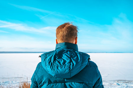 Rear view of man in a winter jacket stands against the background of a frozen river with a winter...