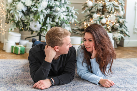 Beautiful young couple man and woman lying on the living room floor next to a nicely decorated Christmas tree and talkの写真素材
